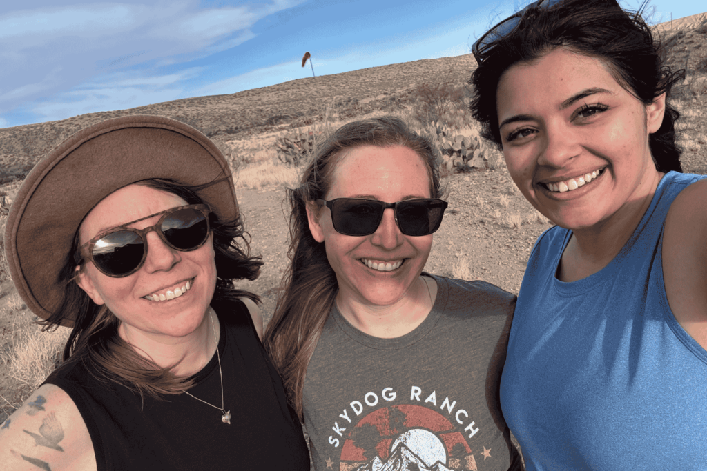 Three women face the camera and smile in the New Mexico high desert with scrub and blue sky behind them. From left to right, Michelle Lute is wearing a black tank top, sunglasses and a hat; in the middle,Mandy Culbertson is wearing a Skydog Sanctuary gray shirt and sunglasses; and on the right, Delani Delgado is wearing a blue tank top and a gold pendant necklace that hits above her breastbone. Michelle has light brown hair, white skin, and a partially visibly tattoo on her right shoulder. Mandy has dark blonde hair, white skin. Delani has medium-toned skin, brown eyes, and black hair. Wildlife for all is a women-led organization and these three are women in conservation.