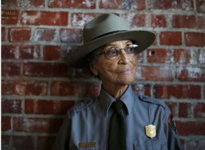 Betty Reid Soskin looks to the right of the image wearing her park ranger uniform as she leans against a red brick wall. She is a trailblazer for women in conservation as we celebrate women's history month.